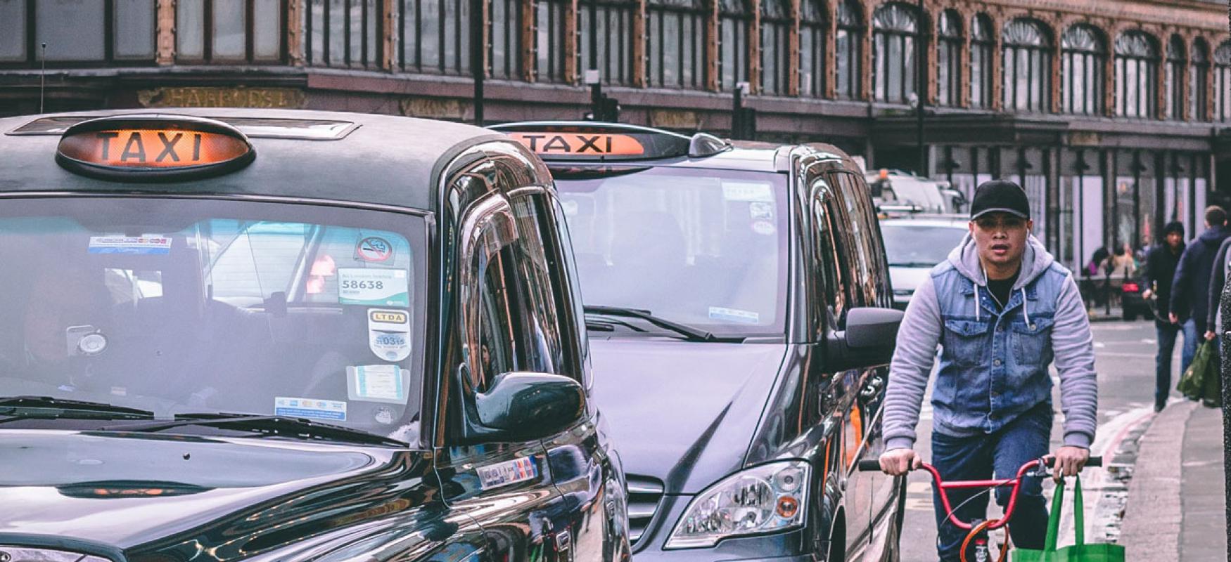 Man riding a bike past two black cabs in London.