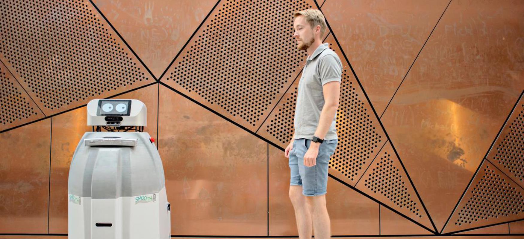 Robot and man standing next to eachother i front of a structured brass wall.