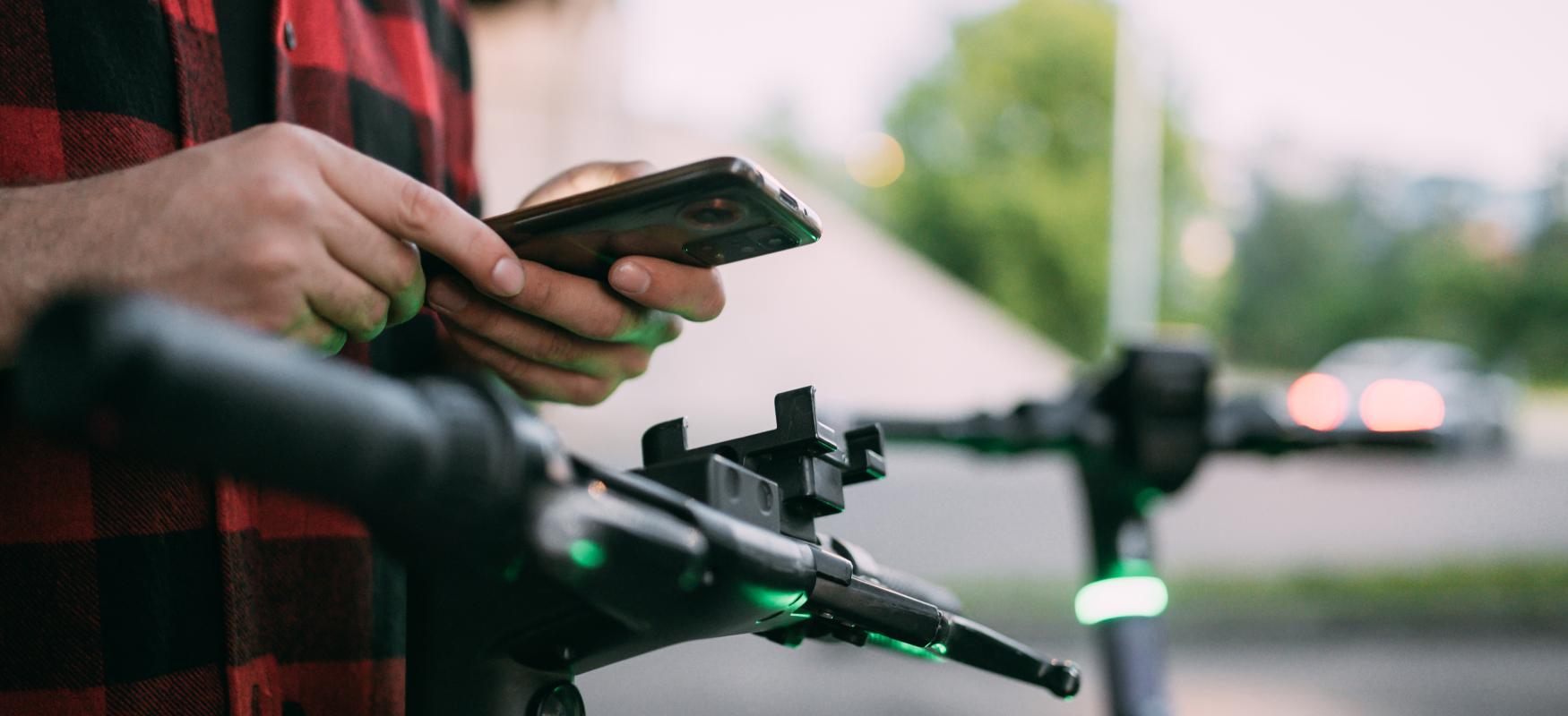 mand står ved sin sparkecykel og kigger på sin smartphone