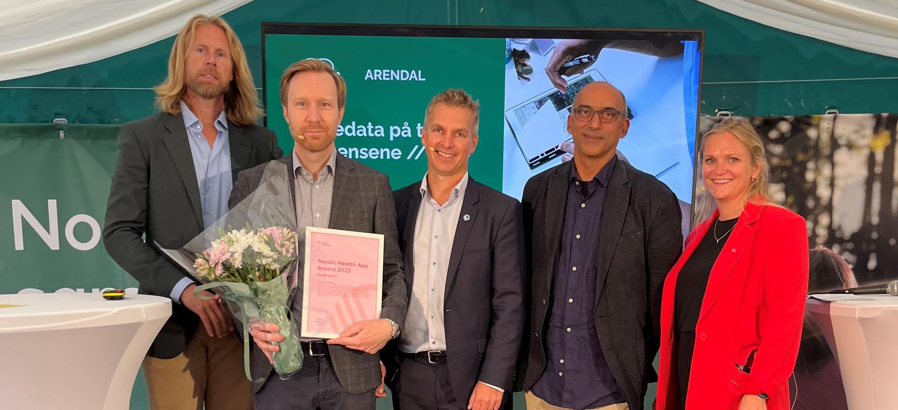 5 persomner står på en scenen. Den ene holder et diplom
