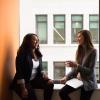 Two young women in blazers sitting and talking, women to the right is sitting with a laptop. Window in the background, looking out onto another building.